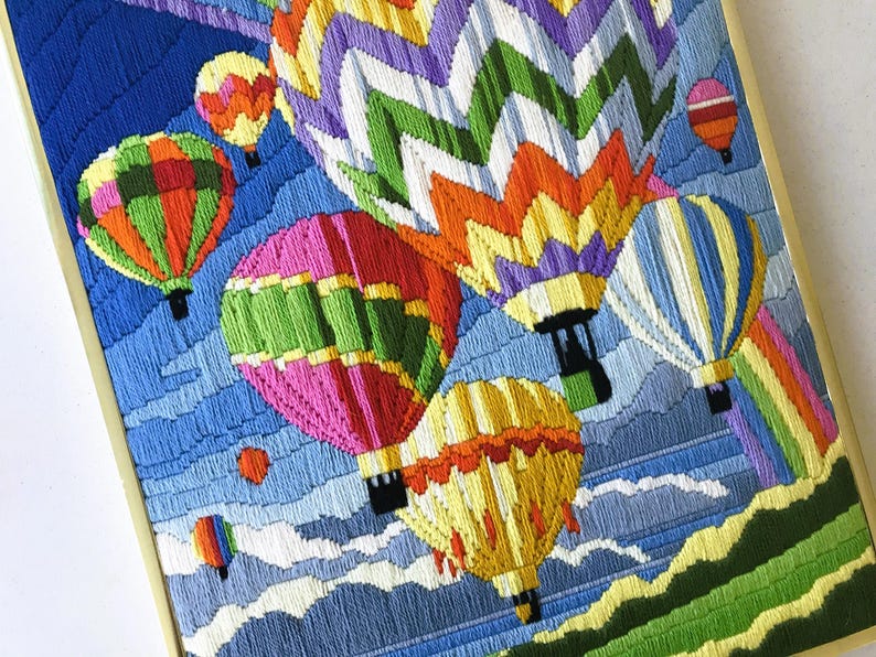 May include: A framed embroidery featuring colorful hot air balloons against a blue sky with clouds. The balloons are various colors, including pink, green, yellow, and orange. The artwork has a detailed, textured appearance.
