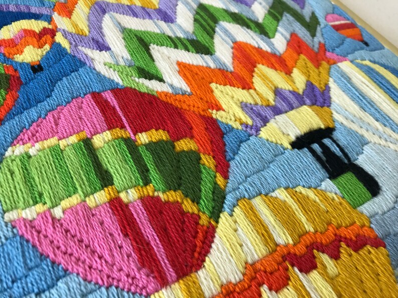 May include: Close-up of a colorful, embroidered artwork featuring hot air balloons. The balloons are in various colors, including pink, green, orange, and yellow, with a blue sky background. The piece is framed.