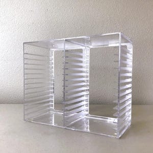 May include: Clear acrylic CD storage unit with multiple compartments. The unit has vertical dividers and horizontal shelves for organizing CDs. The transparent design allows for easy viewing of the contents.