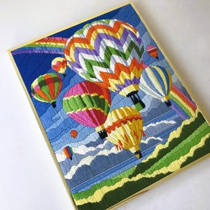 May include: Embroidered artwork featuring colorful hot air balloons floating in a blue sky with a rainbow. The balloons are various colors, including red, green, yellow, and purple. The artwork is framed in a gold-colored frame.