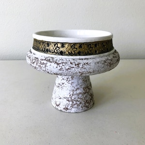 May include: A white pedestal bowl with a textured, speckled surface. The bowl features a gold band with a floral design around the rim. The pedestal base is also white with a textured finish. This decorative bowl is suitable for displaying small items.