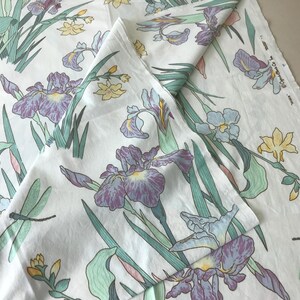 May include: White fabric with a floral pattern featuring purple irises, yellow flowers, green leaves, and dragonflies. The design is repeated across the fabric, creating a botanical theme. The fabric is folded.