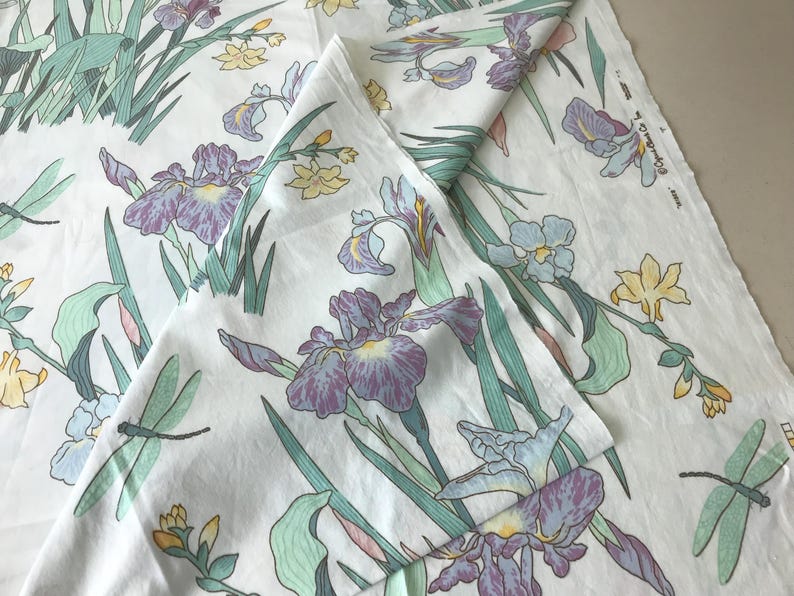 May include: White fabric with a floral pattern featuring purple irises, yellow flowers, green leaves, and dragonflies. The design is repeated across the fabric, creating a botanical theme. The fabric is folded.