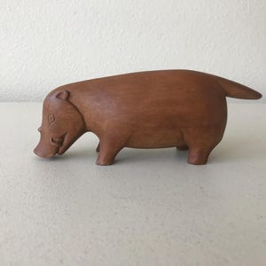 K&ouml;nnte beinhalten: Eine handgeschnitzte Holzskulptur eines Warzenschweins. Die braune Figur hat einen abgerundeten K&ouml;rper, kurze Beine und einen kleinen Schwanz. Das Warzenschwein ist mit einer Schnauze und kleinen Ohren dargestellt, was ihm ein skurriles Aussehen verleiht. Die Skulptur ist ein Dekorationsartikel.