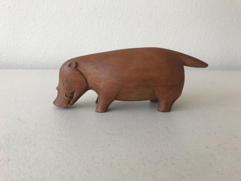 K&ouml;nnte beinhalten: Eine handgeschnitzte Holzskulptur eines Warzenschweins. Die braune Figur hat einen abgerundeten K&ouml;rper, kurze Beine und einen kleinen Schwanz. Das Warzenschwein ist mit einer Schnauze und kleinen Ohren dargestellt, was ihm ein skurriles Aussehen verleiht. Die Skulptur ist ein Dekorationsartikel.