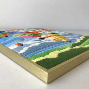 May include: Embroidered artwork featuring colorful hot air balloons floating in a blue sky with white clouds. The artwork is framed in a gold-colored frame, with green and yellow embroidered hills in the foreground.