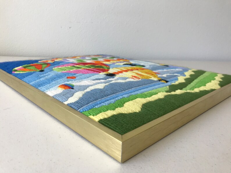 May include: Embroidered artwork featuring colorful hot air balloons floating in a blue sky with white clouds. The artwork is framed in a gold-colored frame, with green and yellow embroidered hills in the foreground.