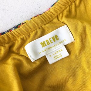 Op de afbeelding: Close-up van een geel kledingstuk met een label met de tekst "Maeve by Anthropologie" en "X-LARGE Made in Vietnam". Het kledingstuk heeft een kleurrijke patroonrand en een gele voering.