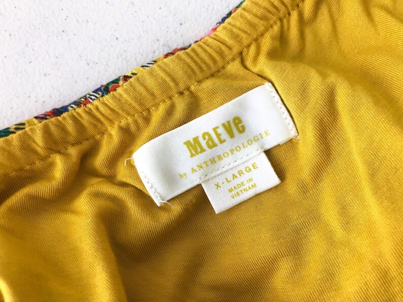 Op de afbeelding: Close-up van een geel kledingstuk met een label met de tekst "Maeve by Anthropologie" en "X-LARGE Made in Vietnam". Het kledingstuk heeft een kleurrijke patroonrand en een gele voering.