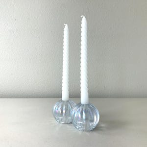 May include: Two white spiral taper candles standing in clear, iridescent glass candle holders. The holders have a rounded, ribbed, pumpkin-like shape with a reflective finish. The candles are tall and slender, featuring a twisted texture throughout.