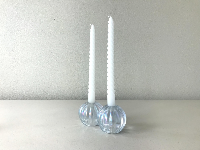 May include: Two white spiral taper candles standing in clear, iridescent glass candle holders. The holders have a rounded, ribbed, pumpkin-like shape with a reflective finish. The candles are tall and slender, featuring a twisted texture throughout.
