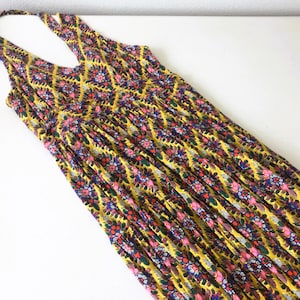 Op de afbeelding: Een gele maxi-jurk met halternek en een bloemenpatroon in roze, blauw en groen. De jurk heeft een V-hals en een geplooid taille. De stof is lichtgewicht en lijkt geschikt voor warm weer.