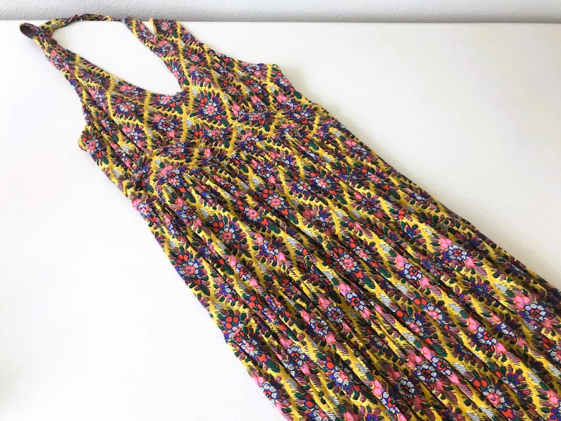 Op de afbeelding: Een gele maxi-jurk met halternek en een bloemenpatroon in roze, blauw en groen. De jurk heeft een V-hals en een geplooid taille. De stof is lichtgewicht en lijkt geschikt voor warm weer.