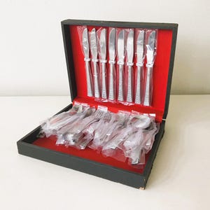 May include: A vintage silverware set in a black box with a red velvet interior. The set includes knives, forks, and spoons, all individually wrapped in plastic. The silverware has a decorative handle design.