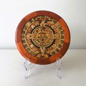 May include: A wooden wall hanging with a circular design featuring a colorful Aztec calendar. The calendar is surrounded by a wooden frame with a red and orange border. The wall hanging is on a clear acrylic stand.