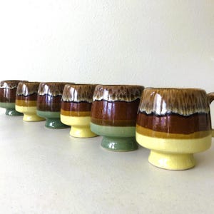 May include: Set of seven brown, green, and yellow ceramic mugs with a glazed finish. The mugs have a unique, wavy design and some have handles.