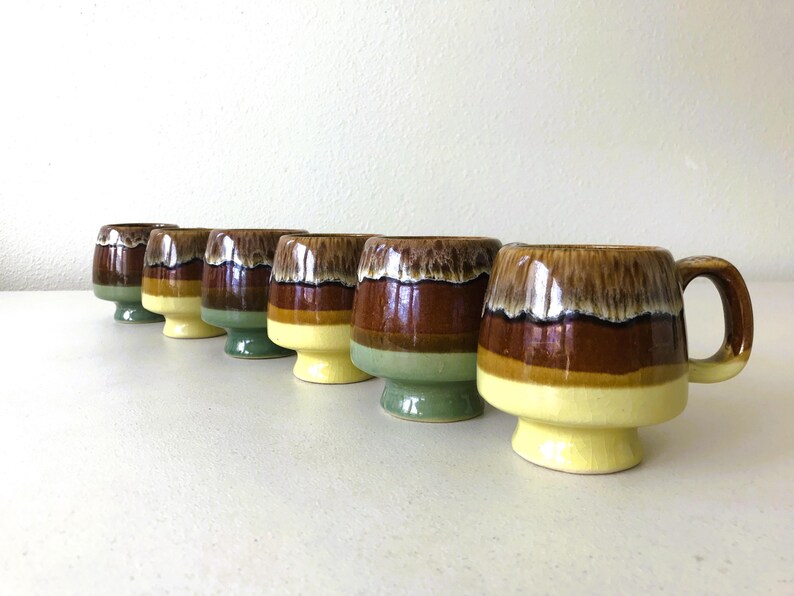 May include: Set of seven brown, green, and yellow ceramic mugs with a glazed finish. The mugs have a unique, wavy design and some have handles.