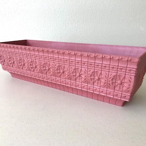 May include: A rectangular pink planter box featuring a raised, repeating floral pattern along the exterior sides. The container has a textured surface with a wide, flat rim and a slightly tapered base, providing a structured, open-top design.