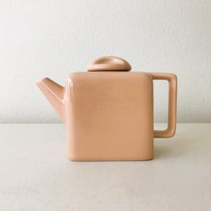 May include: A peach-colored ceramic teapot with a square body, a handle, and a spout. The lid is a rounded shape that sits on top. The teapot is on a white surface against a white background.
