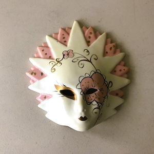 May include: A decorative ceramic mask with a sunburst design. The mask is primarily white with pink and gold accents. It features a floral design on the right side and gold detailing around the eyes and mouth.