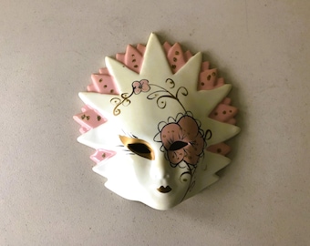 May include: A decorative ceramic mask with a sunburst design. The mask is primarily white with pink and gold accents. It features a floral design on the right side and gold detailing around the eyes and mouth.