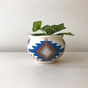 May include: A small, round, off-white ceramic planter with a geometric blue, brown, and pink design. The planter holds a small green plant with textured leaves. The pot has a Southwestern style design.
