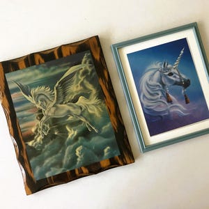 May include: Two framed fantasy art prints. One features a white Pegasus with outstretched wings soaring through clouds, framed in a dark, textured wood. The other is a unicorn portrait with a blue frame, set against a starry background.