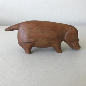 K&ouml;nnte beinhalten: Eine handgeschnitzte Holzskulptur eines stilisierten Wildschweins. Die braune Figur hat eine glatte Oberfl&auml;che mit geschnitzten Details, die die Merkmale des Tieres definieren, einschlie&szlig;lich Schnauze, Ohren und Schwanz. Das Wildschwein wird in einer Laufhaltung dargestellt.