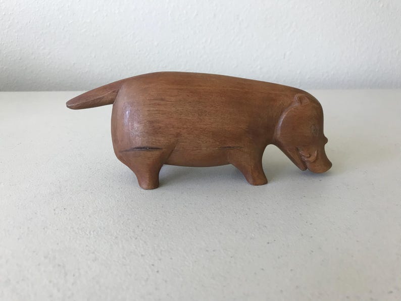 K&ouml;nnte beinhalten: Eine handgeschnitzte Holzskulptur eines stilisierten Wildschweins. Die braune Figur hat eine glatte Oberfl&auml;che mit geschnitzten Details, die die Merkmale des Tieres definieren, einschlie&szlig;lich Schnauze, Ohren und Schwanz. Das Wildschwein wird in einer Laufhaltung dargestellt.