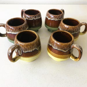 May include: Six brown and yellow ceramic mugs with a glazed finish. The mugs have a rounded shape and a handle on the side.