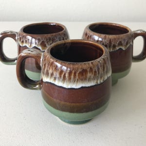 May include: Three brown and green ceramic mugs with a white glaze. The mugs have a rounded shape and a handle on one side.