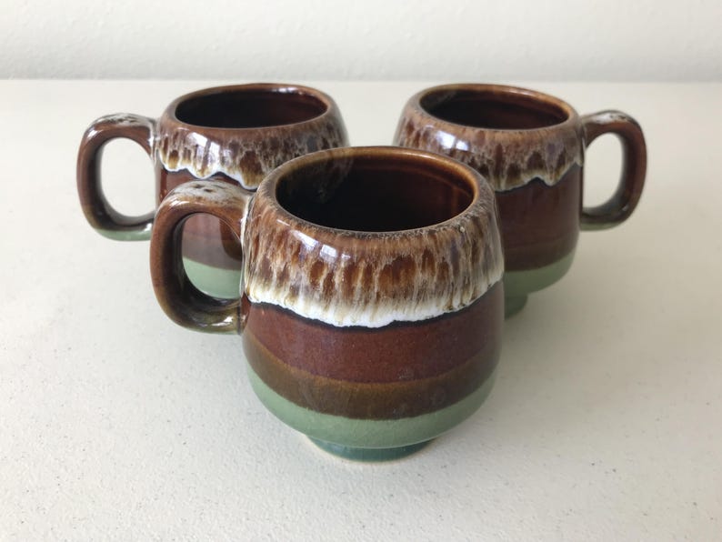 May include: Three brown and green ceramic mugs with a white glaze. The mugs have a rounded shape and a handle on one side.