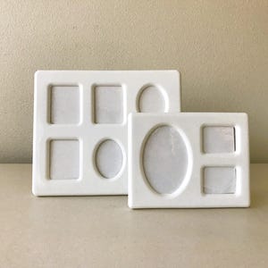 May include: Two white ceramic multi-photo frames. One frame has six openings in square and oval shapes. The other frame has four openings in square and oval shapes. The frames are on a neutral surface.