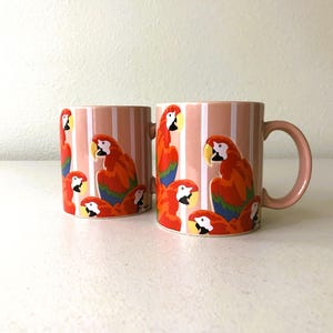 May include: Two ceramic mugs with a pink and white striped pattern. Each mug features a colorful illustration of a parrot with red, orange, yellow, and blue feathers. The mugs have pink handles and are set against a white background.