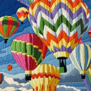 May include: A framed needlepoint artwork featuring several colorful hot air balloons against a blue sky with white clouds. The balloons are various colors, including orange, green, pink, and yellow. The artwork is framed in a gold-colored frame.