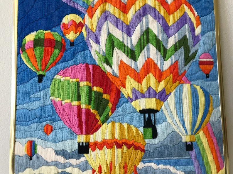 May include: A framed needlepoint artwork featuring several colorful hot air balloons against a blue sky with white clouds. The balloons are various colors, including orange, green, pink, and yellow. The artwork is framed in a gold-colored frame.