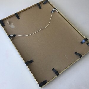 May include: Back of a rectangular picture frame with a gold-colored metal frame. The back is made of cardboard and secured with black clips. A white string is attached for hanging.