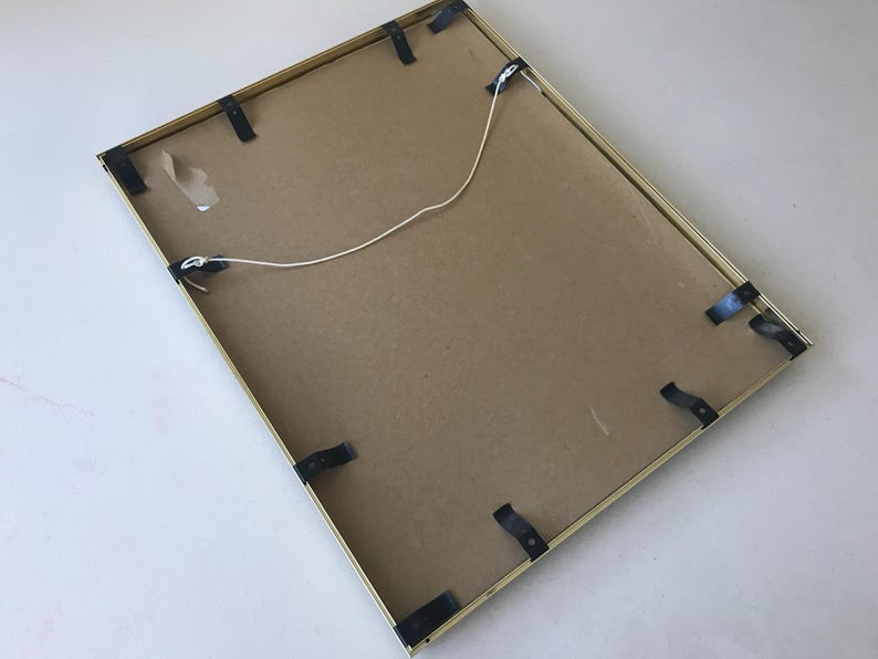 May include: Back of a rectangular picture frame with a gold-colored metal frame. The back is made of cardboard and secured with black clips. A white string is attached for hanging.