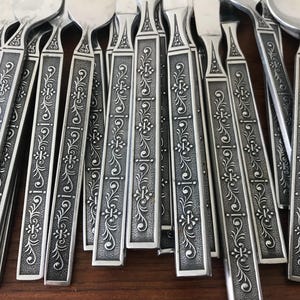 May include: A collection of silver-toned metal flatware pieces featuring a textured, rectangular handle design. Each handle is decorated with a repeating embossed floral and scroll pattern that runs vertically along the center of the stem.
