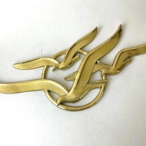 May include: A brass wall sculpture featuring three stylized birds in flight, with a circular element. The birds are depicted with outstretched wings, creating a dynamic and elegant design. The sculpture has a polished, golden finish.