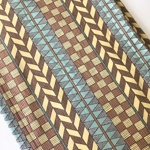 May include: A folded fabric with a repeating geometric pattern. The design features alternating rows of brown, yellow, and light blue shapes, including chevrons, squares, and triangles. The fabric has a textured appearance.