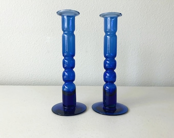 May include: A pair of cobalt blue glass candle holders with a textured, bulbous design. The holders have a wide base and a cylindrical shape.