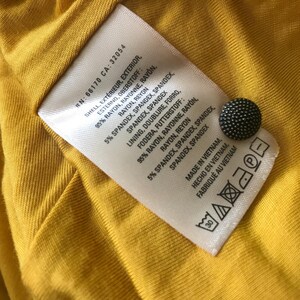 Op de afbeelding: Close-up van een gele stof met een wit label en een zwarte knoop. Het label geeft details over de samenstelling van de stof in het Engels, Frans en Spaans, inclusief rayon en spandex. Het label vermeldt dat het item in Vietnam is gemaakt.