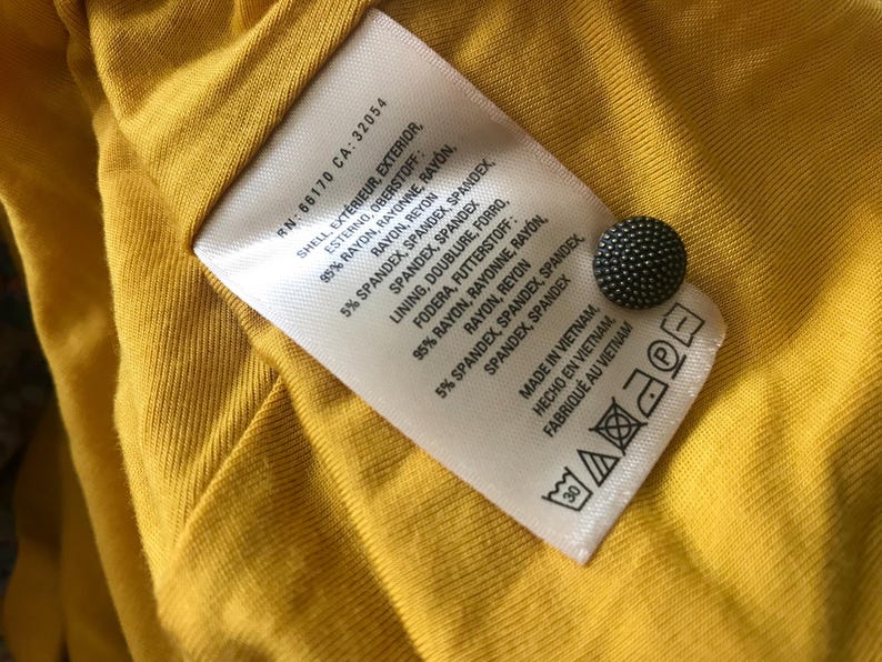 Op de afbeelding: Close-up van een gele stof met een wit label en een zwarte knoop. Het label geeft details over de samenstelling van de stof in het Engels, Frans en Spaans, inclusief rayon en spandex. Het label vermeldt dat het item in Vietnam is gemaakt.