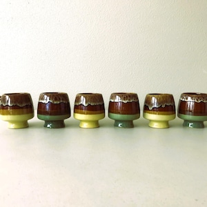 May include: Six brown and green ceramic mugs with a glazed finish. The mugs have a rounded shape and a wide base. One mug has a handle.
