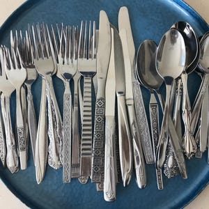 May include: A collection of assorted stainless steel flatware including forks, knives, and spoons with various decorative handle patterns, all arranged together on a round, textured blue metal tray with decorative side handles.