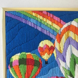 May include: A framed, colorful needlepoint artwork featuring hot air balloons and a rainbow against a blue sky. The balloons are in shades of orange, green, yellow, and pink. The rainbow has stripes of red, orange, yellow, green, blue, and purple.