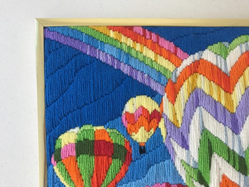 May include: A framed, colorful needlepoint artwork featuring hot air balloons and a rainbow against a blue sky. The balloons are in shades of orange, green, yellow, and pink. The rainbow has stripes of red, orange, yellow, green, blue, and purple.