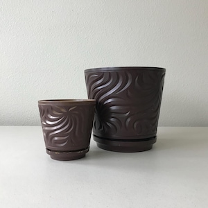 May include: Two brown decorative flower pots with matching saucers. The pots have a swirling, embossed design. The larger pot is approximately 7 inches in diameter and 6 inches tall. The smaller pot is about 4 inches in diameter and 4 inches tall.