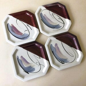 May include: Set of four octagonal ceramic plates. Each plate has a white and burgundy color scheme, with a painted design of blue, purple, and black brushstrokes. The plates are arranged in a square formation.